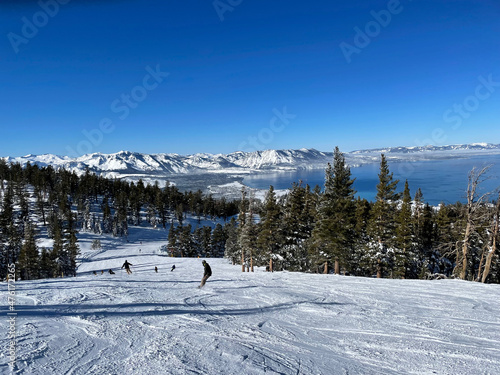 Scenic view of skiers and snowboarders on the slopes of a ski resort on a bluebird winter day, with Lake Tahoe in the background