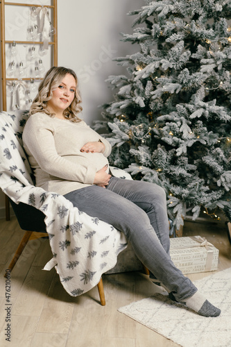 Pregnant Woman sitting chair armchair living room near Christmas tree dressed warm sweater