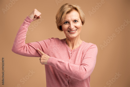 Adorable self confident woman pointing finger on her arm muscle, pumping in gym, discount. Indoor studio shot isolated on beige background