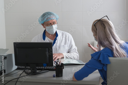 A doctor in a white coat, mask and cap in the office sits at table with a patient and listens to her complaints. Consultative reception . Selective focus