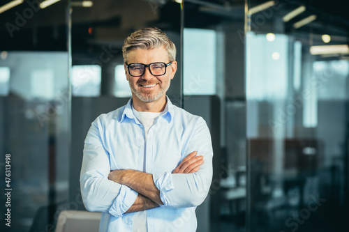 Portrait of senior and experienced male boss boss, with arms crossed, smiling and looking at camera, businessman with glasses, and modern office