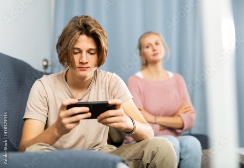 Teenage son sitting on sofa and playing on the phone, while mother trying to talk to him