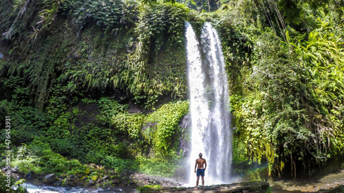 A man in swimsuit standing under two levelled waterfall in Lombok, Indonesia. Tiu Kelep Waterfall is surrounded by lush green plants from each side. Long and powerful waterfall. Beauty of the nature.