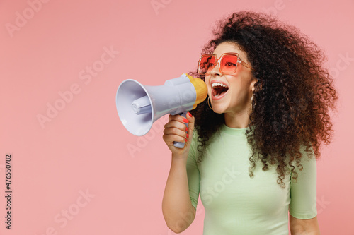 Exultant happy vivid young curly latin woman 20s wear mint t-shirt sunglasses hold scream in megaphone announces discounts sale Hurry up isolated on plain pastel light pink background studio portrait