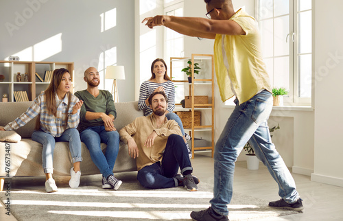 Diverse group of happy people hanging out at home, playing charades and having fun together. Young African man acting out a syllable, word, or phrase to give his friends a clue of what it might be