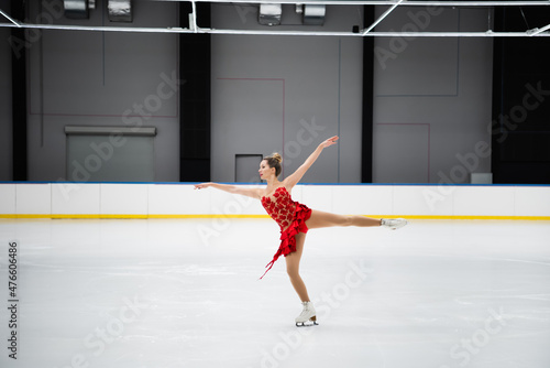 full length of figure skater in red dress performing camel spin in professional ice arena