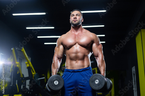 Front view of a muscular bodybuilder posing with dumbbells in blue shorts in a gym