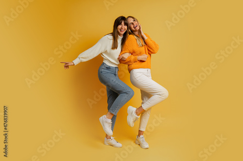 Two pretty women , best friends in stylish autumn casual clothes having fun over yellow background in studio. Full lenght.