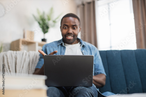 Happy african american man in casual clothes sitting on couch and using wireless laptop. Young guy browsing internet during leisure time at cozy home.