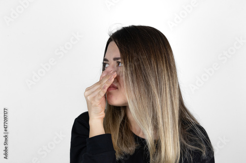 Displeased woman covers nose with hand, smells something awful, pinches nose, frowns in displeasure, sees pile of garbage, dressed in casual clothes, isolated on white background. Side profile photo.