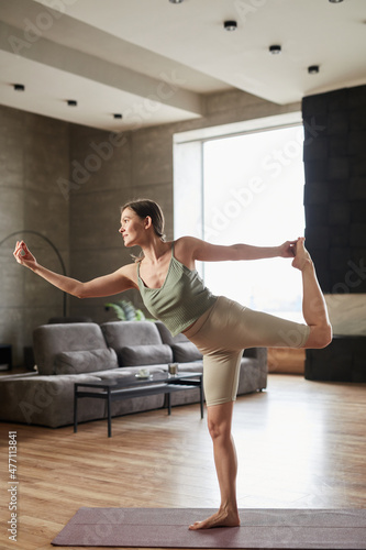 Young slim woman standing on exercise mat and stretching her arms and legs during training in the morning in the room