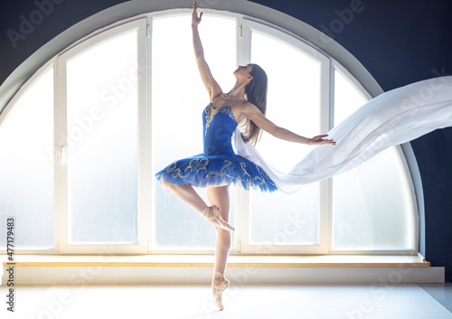 Focused young ballerina dressed in blue tutu costume makes pirouette at ballet studio in front of big round shaped window background in natural ambient daylight atmosphere