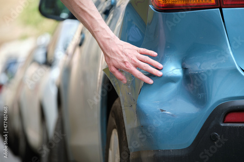 Driver hand examining dented car with damaged fender parked on city street side. Road safety and vehicle insurance concept