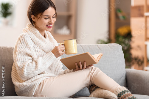 Beautiful young woman drinking tasty tea and reading book on sofa at home