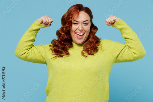 Young strong fun chubby overweight plus size big fat fit woman in green sweater showing biceps muscles on hand demonstrating strength power isolated on plain blue background People lifestyle concept