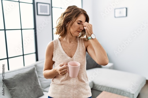 Middle age woman drinking a cup coffee at home tired rubbing nose and eyes feeling fatigue and headache. stress and frustration concept.