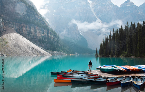 Tiny person standing on dock with colourful canoes at turquoise blue lake surrounded by mountains