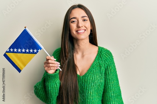 Young hispanic girl holding bosnia herzegovina flag looking positive and happy standing and smiling with a confident smile showing teeth