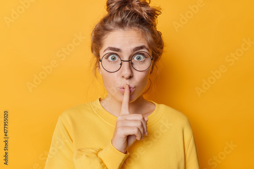 Stunned emotional beautiful woman hushing with index finger shares secret makes taboo gesture stares through round spectacles wears casual jumper isolated over yellow background. Shh be quiet