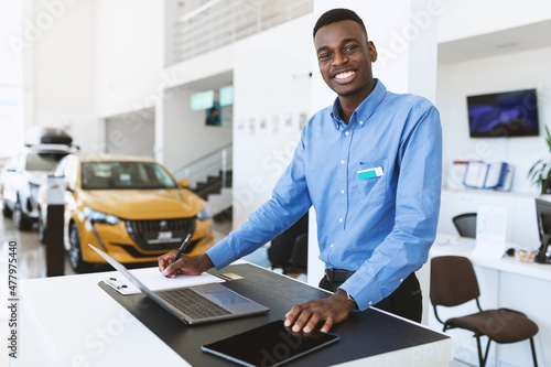 Portrait of happy car salesman standing at work desk, smiling at camera, using laptop and touch pad in auto dealership