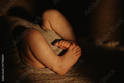 The legs of a newborn baby in a bodysuit who is lying on a sofa in the sunlight