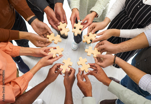 Business and cooperation. Multiracial people stand in circle and connect wooden pieces of puzzles they hold in their hands. Cropped image of hands holding puzzle pieces symbolizing unity and teamwork.