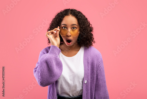 Emotional young African American lady touching sunglasses and looking at camera in shock over pink studio background