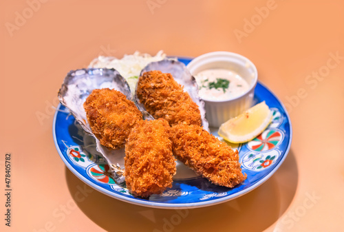 Japanese shellfish dish of battered and deep fried oysters made by adding egg whites and bread crumbs called kaki-furai or kaki fry served in their shell with a sauce and a lemon slice in a plate.