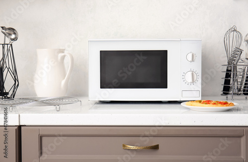 Modern microwave oven and plate with pizza on counter near light wall