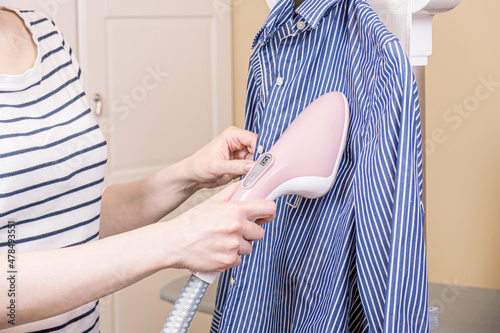 Woman ironing a striped business shirt with a handheld garment steamer. Using a household steamer hand ironing machine to steam clean clothes. Household chores.