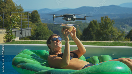 CLOSE UP: Food delivery service drone brings a bowl to a woman relaxing in pool.