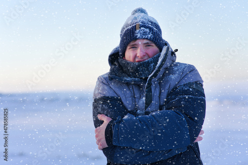 Portrait of frozen suffering guy, young handsome freezing man standing walking outdoors at winter snowy cold frosty day, shaking, trembling, shivering because of extreme low temperature in jacket, hat