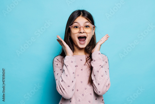 Portrait of desperate and shocked little girl wearing blue T-shirt over blue background holding hands near face, with mouth wide open.