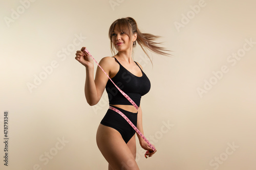 Slim healthy girl in black underwear with measuring tape at waist. Fit Woman poses in high waist panties and top. Perfect female body. Fitness, Diet or body care concept. Female shows her flat belly