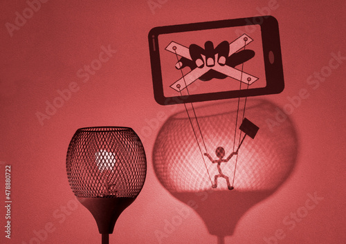 Gaslight with silhouette of puppet on strings being manipulated by a hand in mobile phone cast by the lamp on wall, social media gaslighting concept illustration