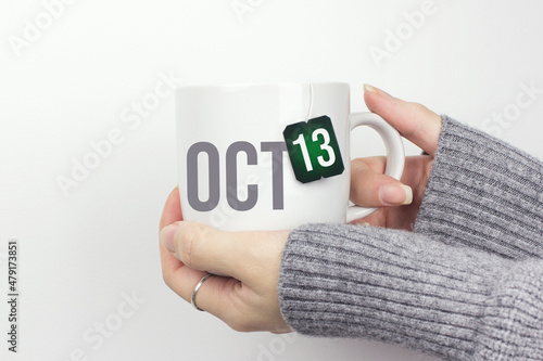 October 13rd. Day 13 of month, Calendar date. Closeup of female hands in grey sweater holding cup of tea with month and calendar date on teabag label. Autumn month, day of the year concept.