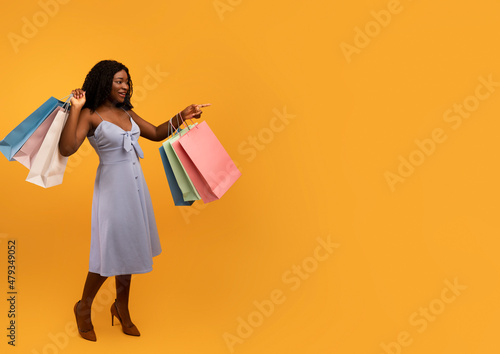 Young black woman in dress holding bright shopping bags, pointing at empty space, offering place for your ad