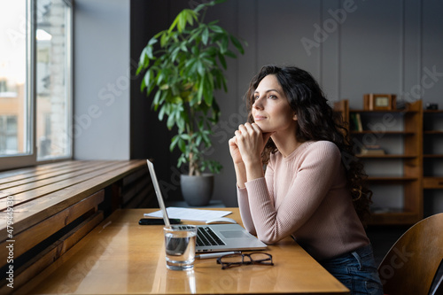 Young dreamy smiling female entrepreneur thinking about business goals and objectives, enjoying good window view while working in office, pleased businesswoman resting from computer work