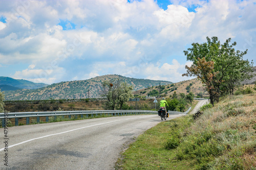 A traveler cyclist rides on an asphalt road among the Crimean mountains and vineyards.