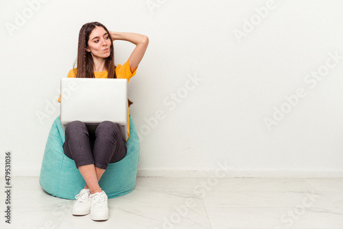 Young caucasian woman working with laptop sitting on a puff isolated on white background touching back of head, thinking and making a choice.