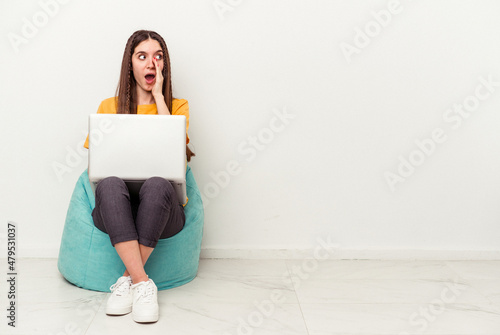Young caucasian woman working with laptop sitting on a puff isolated on white background is saying a secret hot braking news and looking aside