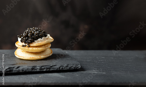 A stack of blini pancakes with black caviar and sour cream on a slate, copy space