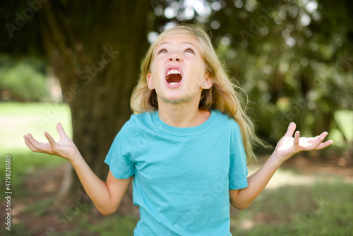 Caucasian little kid girl wearing blue T-shirt standing outdoors crazy and mad shouting and yelling with aggressive expression and arms raised. Frustration concept.