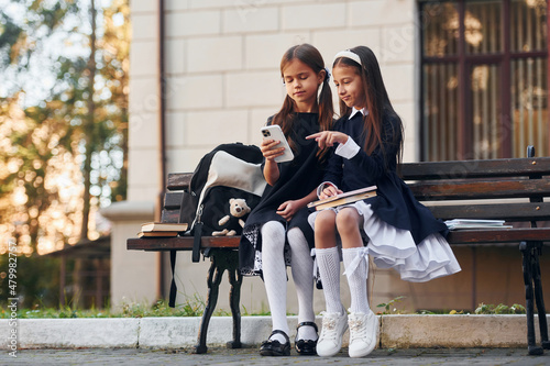 Two schoolgirls is sitting outside together near school building