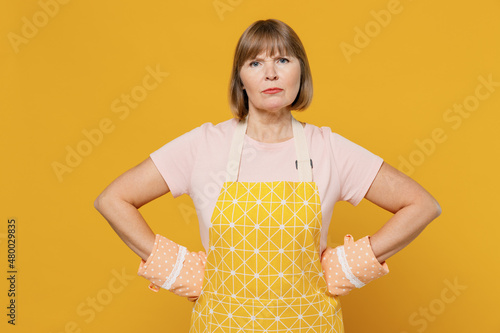 Elderly strict angry indignant sad cooker housekeeper housewife woman 50s in orange apron oven mitt stand akimbo isolated plain on yellow background studio portrait People household lifestyle concept
