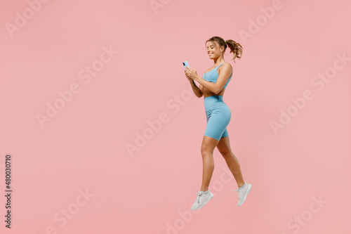 Full body young sporty athletic fitness trainer woman wear blue tracksuit spend time in home gym jump high use mobile cell phone isolated on pastel plain light pink background. Workout sport concept.