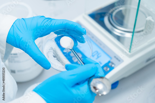 Digital analytical balance for accurate weighing of samples. Close up scientist hands in rubber gloves use metal spatula for weighing white powder of the substance.