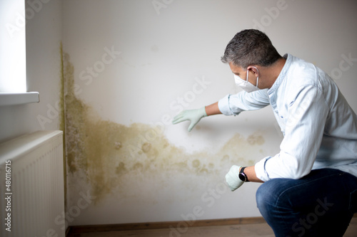 Man with mouth nose mask and blue shirt and gloves n front of white wall with mold