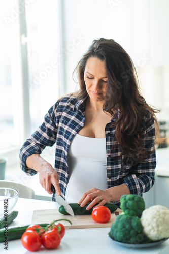  pregnant woman cook salad cutting cucumber on wooden cutting board with fresh vegetables and fruit on table in kitchen. Pregnancy, maternity, healthy lifestyle concept.