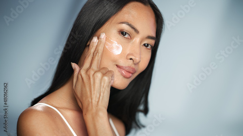 Female Beauty Portrait. Beautiful Asian Brunette Woman Touching Natural, Healthy Skin, Using Facial Moisturizer, Applying Face Cream. Wellness and Skincare Concept on Isolated Background.